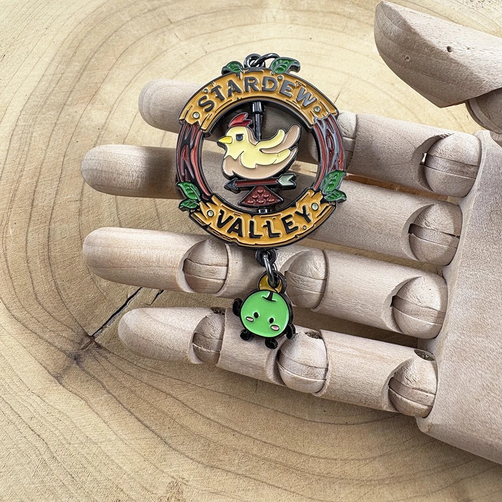 Chaveiro da placa de Stardew Valley, com galinha e Junimo, na mão de um manequim de madeira