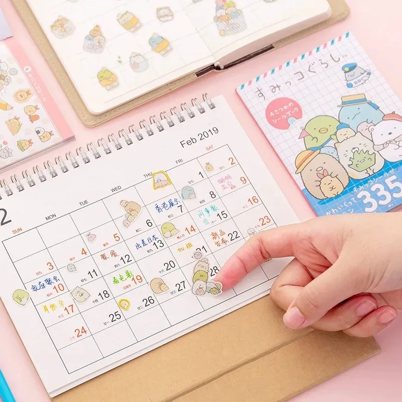 Mão aplicando adesivos de Sumikko Gurashi num calendário de mesa rosa, ao lado de outro livro de adesivos