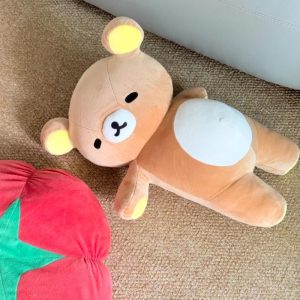 Rilakkuma deitado sobre um tapete, urso marrom de barriga clara e orelhas amarelas